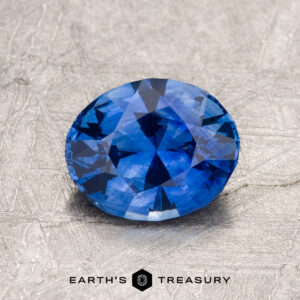 1.56-Carat Ceylon Sapphire (Heated), oval-cut, showcased on a textured gray surface with the “EARTH’S TREASURY” logo and text displayed beneath the gemstone.