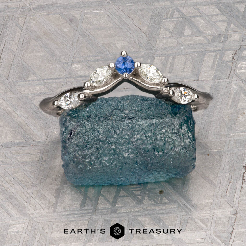 The "Multiflora" band in platinum, featuring three clear marquise stones and a blue pear-shaped stone, stands upright on a rough blue crystal with the "Earth's Treasury" logo below.