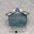 The "Multiflora" band in platinum, featuring three clear marquise stones and a blue pear-shaped stone, stands upright on a rough blue crystal with the "Earth's Treasury" logo below.