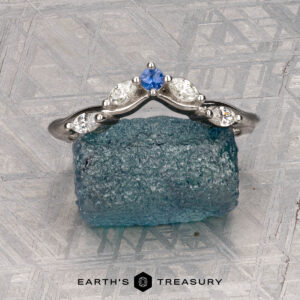 The "Multiflora" band in platinum, featuring three clear marquise stones and a blue pear-shaped stone, stands upright on a rough blue crystal with the "Earth's Treasury" logo below.