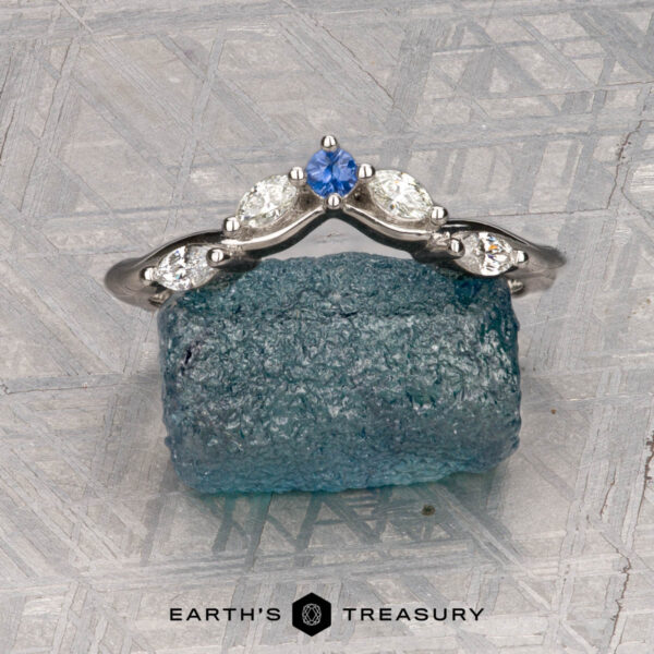 The "Multiflora" band in platinum The "Multiflora" band in platinum, featuring three clear marquise stones and a blue pear-shaped stone, stands upright on a rough blue crystal with the "Earth's Treasury" logo below.