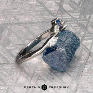 The "Multiflora" band in platinum, featuring three clear marquise stones and a blue pear-shaped stone, stands upright on a rough blue crystal with the "Earth's Treasury" logo below.