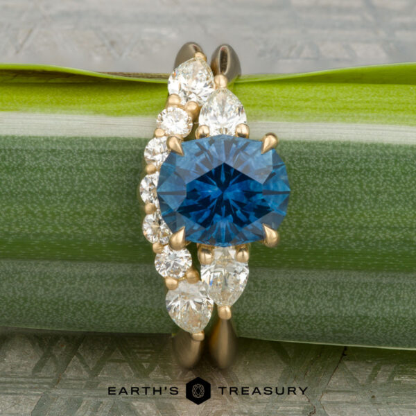The "Multiflora" ring in 18K yellow gold with 1.71-carat Montana sapphire, aloingside the "Clematis" band in 18K yellow gold The "Multiflora" ring in 18K yellow gold with a 1.71-carat Montana sapphire is paired with the "Clematis" band, both from Earth's Treasury. Displayed atop a green leaf, six round white diamonds evoke Clematis petals. Logo at the bottom.