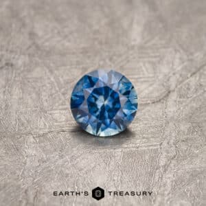 A close-up image showcases a radiant 0.84-carat medium blue Montana sapphire, expertly round-cut and placed on a textured gray surface. The gemstone dazzles with an array of blue shades, while the text "EARTH'S TREASURY" is elegantly displayed at the bottom center.