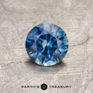 A 0.84-carat heated Montana Sapphire, round brilliant-cut, displayed on a textured metallic surface with "EARTH'S TREASURY" at the bottom.