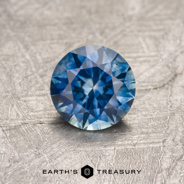0.84-Carat Montana Sapphire (Heated) A 0.84-carat heated Montana Sapphire, round brilliant-cut, displayed on a textured metallic surface with "EARTH'S TREASURY" at the bottom.