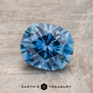 A 1.90-carat Medium Blue Montana Sapphire (heated) rests on a textured wooden surface, with "Earth's Treasury" branding below.
