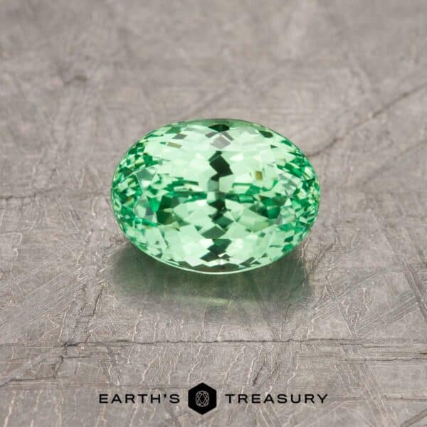 A 4.43-carat light green Tsavorite garnet with an oval cut sits on a textured gray surface. The gem exhibits sparkling facets and reflections. Text at the bottom reads "Earth's Treasury".