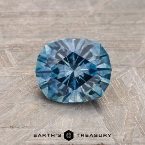 A 1.26-carat teal blue heated Montana Sapphire is showcased on a textured gray surface, with "Earth's Treasury" displayed at the bottom.