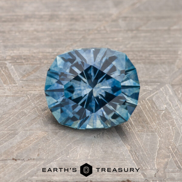 1.26-Carat Teal Blue Montana Sapphire (Heated) A 1.26-carat teal blue heated Montana Sapphire is showcased on a textured gray surface, with "Earth's Treasury" displayed at the bottom.