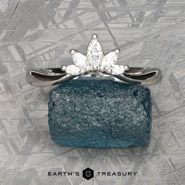 The "Vilaine" band in platinum The "Vilaine" band in platinum with five white gemstones rests on a rough blue stone atop a textured gray surface, with the "Earth's Treasury" logo at the bottom.