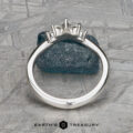 The "Vilaine" band in platinum, with a three-stone setting, stands upright against blue textured stone, exuding bold elegance. The Earth’s Treasury logo appears at the bottom.