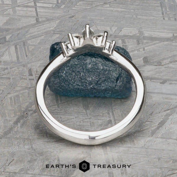 The "Vilaine" band in platinum The "Vilaine" band in platinum, with a three-stone setting, stands upright against blue textured stone, exuding bold elegance. The Earth’s Treasury logo appears at the bottom.