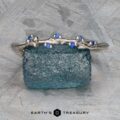 A bracelet in platinum enhanced with blue gemstones is placed on a textured blue background, capturing the calming rhythms associated with the music of the Juniper Band. Below it, the name "Earth's Treasury" is gracefully displayed, reflecting the harmonious spirit of Juniper music.