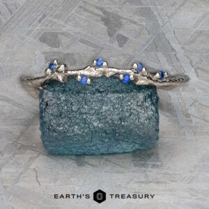 A bracelet in platinum enhanced with blue gemstones is placed on a textured blue background, capturing the calming rhythms associated with the music of the Juniper Band. Below it, the name "Earth's Treasury" is gracefully displayed, reflecting the harmonious spirit of Juniper music.