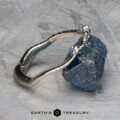 A platinum bracelet from the "Juniper" band collection, embellished with small blue stones, is elegantly displayed on a textured grey surface. The background showcases subtle geometric patterns, reflecting the intricate harmonies associated with the beloved style of Juniper Band.
