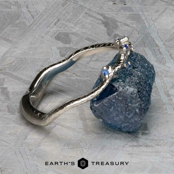 The "Juniper" band in platinum A platinum bracelet from the "Juniper" band collection, embellished with small blue stones, is elegantly displayed on a textured grey surface. The background showcases subtle geometric patterns, reflecting the intricate harmonies associated with the beloved style of Juniper Band.