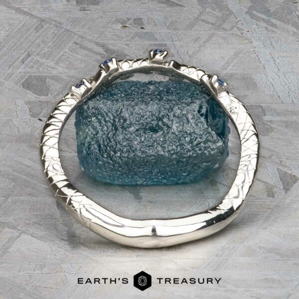 The "Juniper" band in platinum The "Juniper" band in platinum, featuring a textured design with blue gemstones, rests elegantly on a rough blue stone surface, set against a soft gray background evocative of the soothing notes of music.