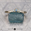 The Marseille Band in 14K yellow gold features a central light blue gemstone and small clear side stones, inspired by elegance. Displayed on a rough blue stone, with "Earth's Treasury" below.