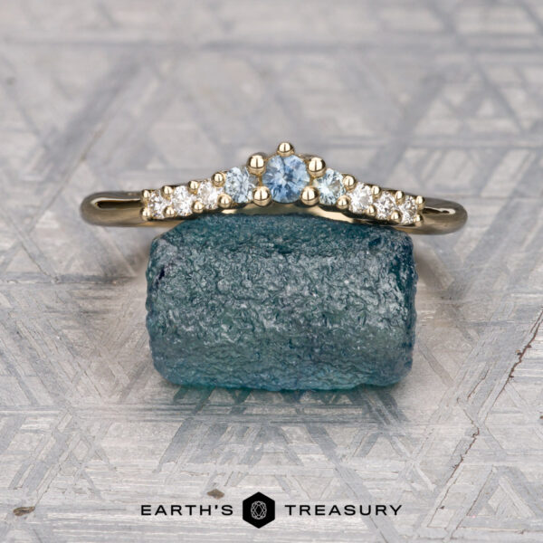 The Marseille band in 14K yellow gold The Marseille Band in 14K yellow gold features a central light blue gemstone and small clear side stones, inspired by elegance. Displayed on a rough blue stone, with "Earth's Treasury" below.