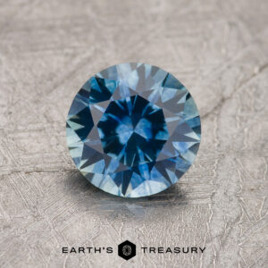 1.21-carat teal blue Montana sapphire (heated), round brilliant-cut, shown on a textured gray surface with "EARTH'S TREASURY" at the bottom.