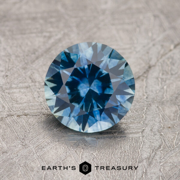 1.21-Carat Teal Blue Montana Sapphire (Heated) 1.21-carat teal blue Montana sapphire (heated), round brilliant-cut, shown on a textured gray surface with "EARTH'S TREASURY" at the bottom.
