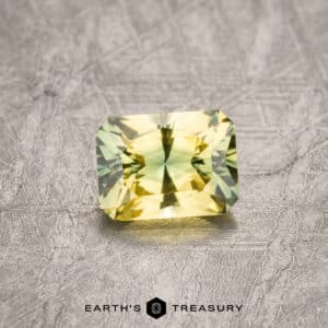 A rectangular, sparkling 1.83-carat Australian sapphire with a vibrant mix of yellow and green hues, placed on a textured grey surface. The faceted gemstone brilliantly showcases its colors and is labeled "EARTH'S TREASURY" below.