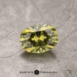A faceted greenish-yellow Australian sapphire lies on a textured, silver-gray surface. The 1.80-carat gem has a complex, symmetrical cut, displaying multiple reflective facets. The text "EARTH'S TREASURY" appears at the bottom, next to a small hexagonal logo.

