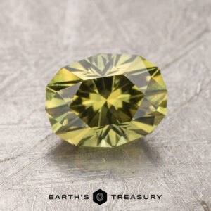 A faceted greenish-yellow Australian sapphire lies on a textured, silver-gray surface. The 1.80-carat gem has a complex, symmetrical cut, displaying multiple reflective facets. The text "EARTH'S TREASURY" appears at the bottom, next to a small hexagonal logo.