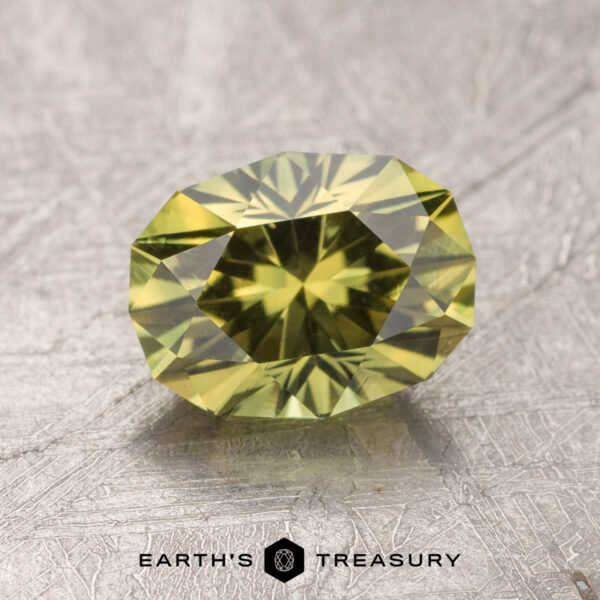 1.80-Carat Australian Sapphire A faceted greenish-yellow Australian sapphire lies on a textured, silver-gray surface. The 1.80-carat gem has a complex, symmetrical cut, displaying multiple reflective facets. The text "EARTH'S TREASURY" appears at the bottom, next to a small hexagonal logo.