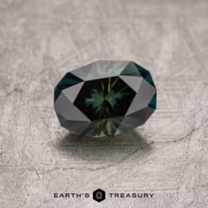 A faceted, oval-shaped 1.73-carat Midnight Blue-Olive particolored Australian sapphire (heated) is displayed on a textured gray surface. The light reflects off its numerous facets, creating a captivating play of colors. The bottom of the image reads "Earth's Treasury.
