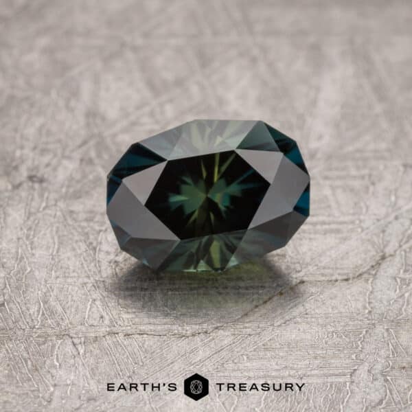 1.73-Carat Australian Sapphire A faceted, oval-shaped 1.73-carat Midnight Blue-Olive particolored Australian sapphire (heated) is displayed on a textured gray surface. The light reflects off its numerous facets, creating a captivating play of colors. The bottom of the image reads "Earth's Treasury.