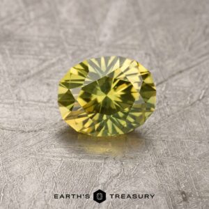 Close-up of a faceted, oval-shaped 1.71-carat greenish-yellow Australian sapphire, displayed on a textured metallic surface. Underneath the gemstone, the text "EARTH'S TREASURY" and its logo are visible.