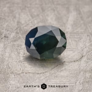 A close-up of a faceted, dark green gemstone with geometric cuts, placed on a textured gray surface. The text "1.52-Carat Midnight Blue-Green Australian Sapphire (Heated)" is visible at the bottom of the image.