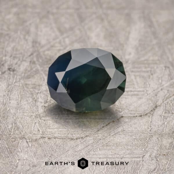 1.52-Carat Australian Sapphire A close-up of a faceted, dark green gemstone with geometric cuts, placed on a textured gray surface. The text "1.52-Carat Midnight Blue-Green Australian Sapphire (Heated)" is visible at the bottom of the image.