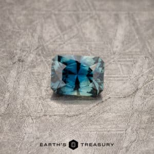 A faceted 0.88-carat teal blue Australian sapphire rests on a textured gray surface. The gem catches the light, displaying intricate cuts and reflecting shades of blue and green. "Earth's Treasury" is written below the gem.