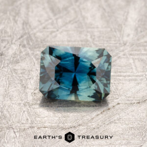 A faceted 0.88-carat teal blue Australian sapphire rests on a textured gray surface. The gem catches the light, displaying intricate cuts and reflecting shades of blue and green. "Earth's Treasury" is written below the gem.