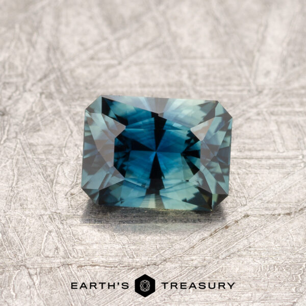 0.88-Carat Australian Sapphire A faceted 0.88-carat teal blue Australian sapphire rests on a textured gray surface. The gem catches the light, displaying intricate cuts and reflecting shades of blue and green. "Earth's Treasury" is written below the gem.