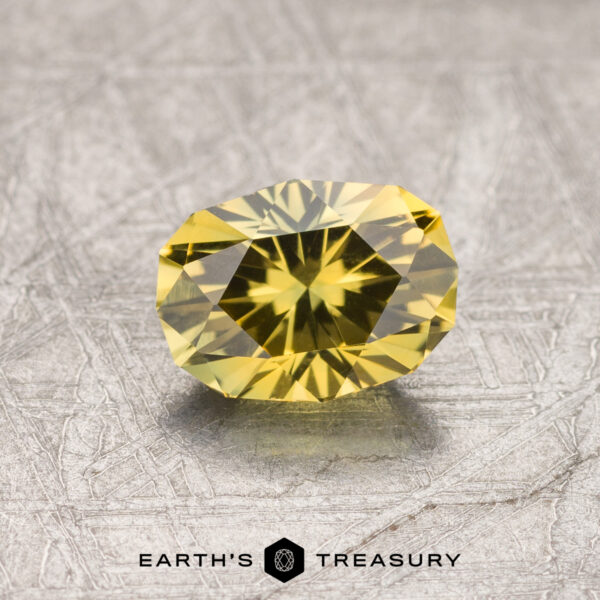 0.96-Carat Australian Sapphire A 0.96-carat golden yellow Australian sapphire, heated for enhanced color, is shown on a textured gray surface. The gemstone is cut into a round brilliant shape, displaying intricate facets that catch the light. The logo "Earth's Treasury" is visible at the bottom of the image.