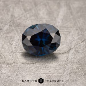A 1.43-carat Midnight Blue Australian Sapphire (heated) with intricate facets lies on a textured gray surface. The gemstone exhibits a star-like pattern and is positioned to reflect light beautifully. The branding "Earth's Treasury" appears beneath the stone.