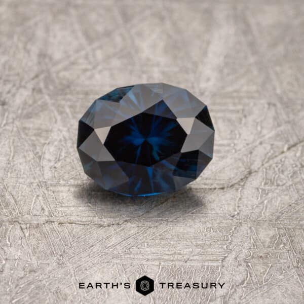 1.43-Carat Australian Sapphire A 1.43-carat Midnight Blue Australian Sapphire (heated) with intricate facets lies on a textured gray surface. The gemstone exhibits a star-like pattern and is positioned to reflect light beautifully. The branding "Earth's Treasury" appears beneath the stone.