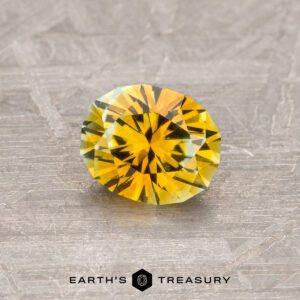 A 0.83-carat golden yellow Montana sapphire, heated and cut in a round brilliant shape, rests on a textured gray surface. The gem displays vibrant facets of yellow and green, reflecting light. The text "Earth's Treasury" is visible at the bottom of the image.