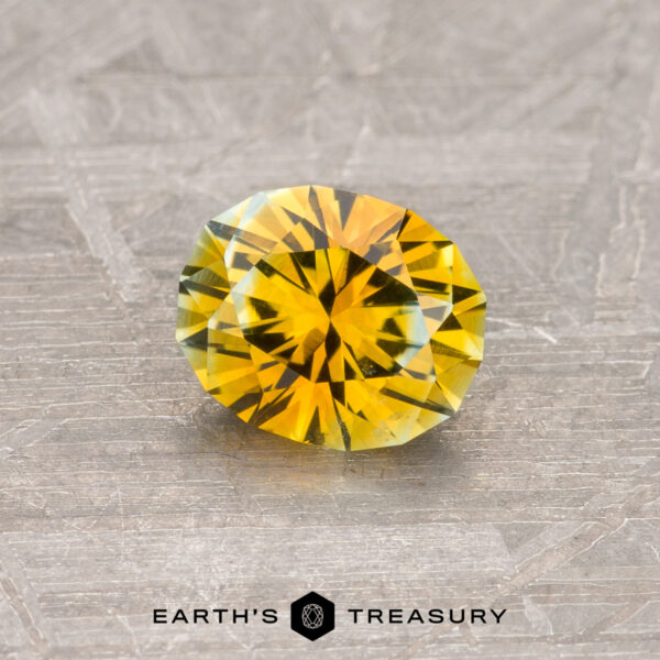 0.83-Carat Montana Sapphire (Heated) A 0.83-carat golden yellow Montana sapphire, heated and cut in a round brilliant shape, rests on a textured gray surface. The gem displays vibrant facets of yellow and green, reflecting light. The text "Earth's Treasury" is visible at the bottom of the image.