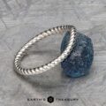 A twisted platinum "Winnie" band rests on a rough blue gemstone, displayed on a textured gray surface with the text "EARTH'S TREASURY" at the bottom.