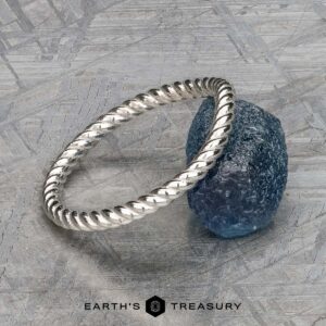 A twisted platinum "Winnie" band rests on a rough blue gemstone, displayed on a textured gray surface with the text "EARTH'S TREASURY" at the bottom.