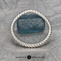 A twisted platinum "Winnie" band bracelet stands upright in front of a rough blue gemstone on a textured metallic surface, with the text "EARTH'S TREASURY" below.
