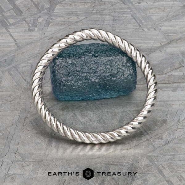 The "Winnie" band in platinum A twisted platinum "Winnie" band bracelet stands upright in front of a rough blue gemstone on a textured metallic surface, with the text "EARTH'S TREASURY" below.