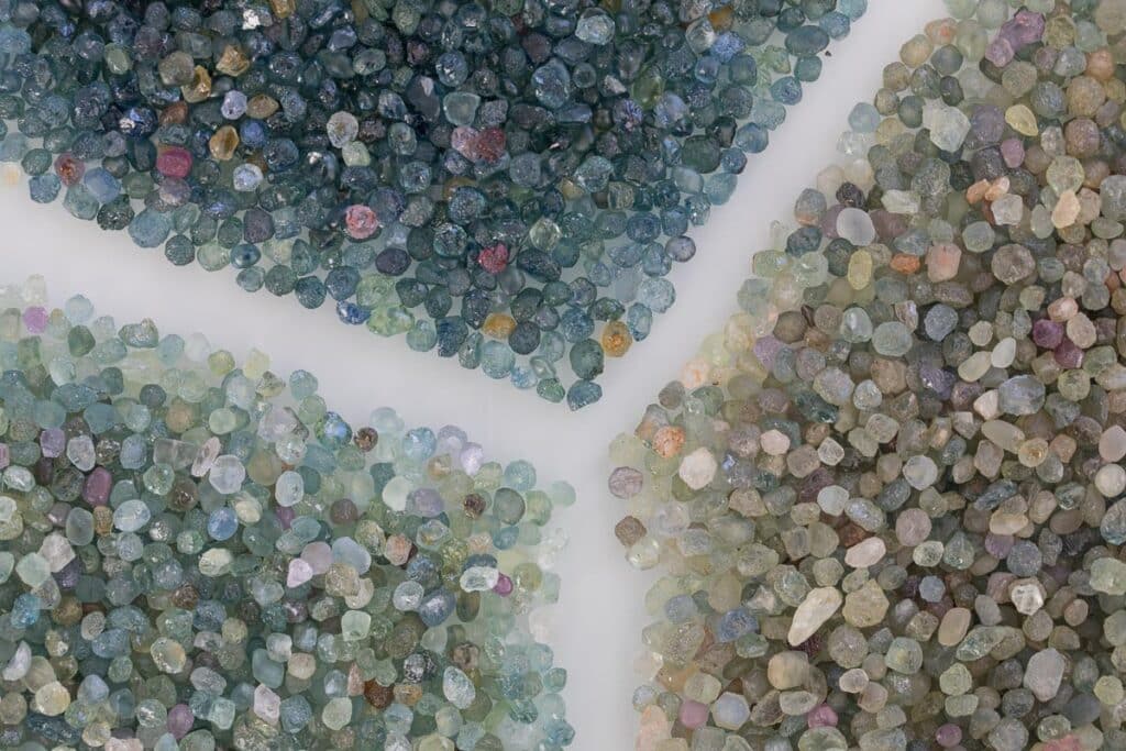 The product _B3A4889-1200-BW content features an array of small, multicolored mineral stones, including exquisite Montana Sapphires, meticulously arranged into quadrants on a light surface to highlight their diverse hues and textures.
