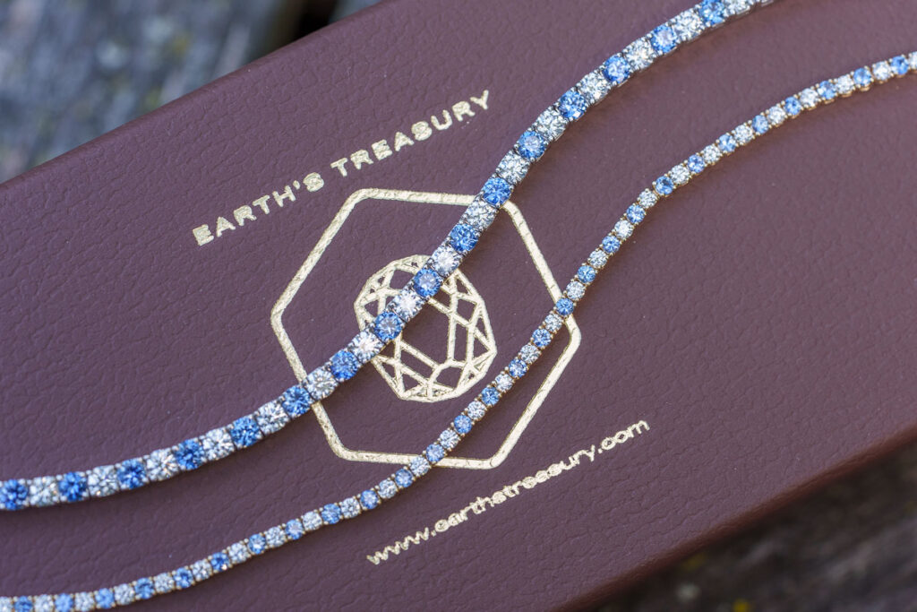The _DSC0865-1200-BW showcases two gemstone tennis bracelets with alternating blue Montana sapphires and clear stones on a brown “Earth’s Treasury” box, highlighting exquisite craftsmanship.