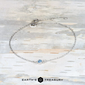 A delicate bracelet crafted from 14k white gold, featuring a luminous Montana sapphire centerpiece, elegantly displayed on a light wooden surface. The bottom proudly showcases the brand name "Earth's Treasury.